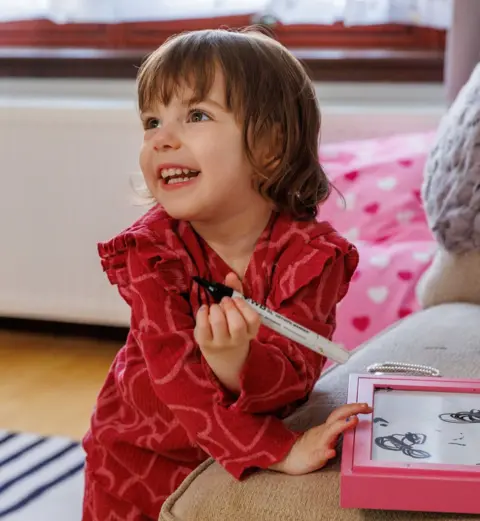 PA Media A young girl in a red outfit playing with an etch o sketch type toy in a living room