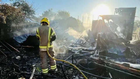 DWFRS A firefighter hoses down a mangled pile of charred material against a low afternoon sun.