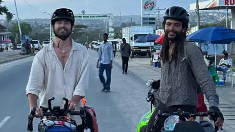 Contributed Two men sit on bikes on the side of a road. They both wear dark helmets and loose shirts. Behind them there are vehicles and people milling about. 