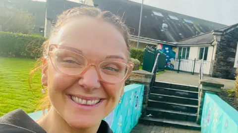 Gráinne Joughin A lady with her hair tied back and wearing pink-coloured, transparent glasses and earrings smiles at the camera. She is standing outside in front a set of steps leading to the patio area of a building.