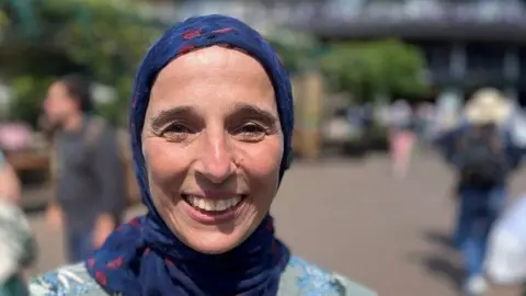 Carmen Torres has a blue headscarf and she is wearing a pale blue top. People are walking around behind her.