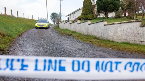 A police car sits at the top of a gravel laneway. In the background is a house, in the foreground is blue and white tape with 'police line do not cross' written on it. 