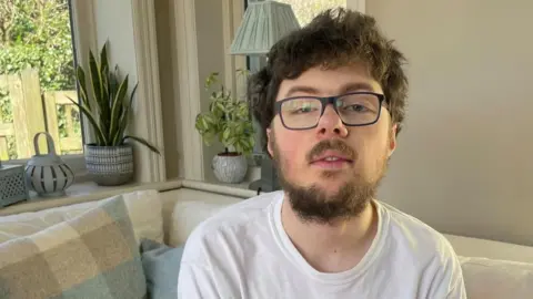 Young man with brown hair and a beard sits on a sofa, by a window. Plants and a lamp are on the windowsill, with fencing and a hedge visible outside. He is wearing spectacles and a white top.