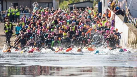 Dozens of people wearing dry suits, wet suits and colourful swimming caps run into the sea on a sunny day for the Kessock Ferry Swim.