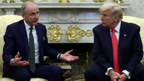 Reuters Micheál Martin sits in the Oval Office in a chair opposite Donald Trump. Martin is speaking and has his arms open. Trump is sat with his hands held and is looking at Martin.