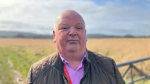 A man in a pink shirt and quilted jacket standing a the gate at the entrance to a field