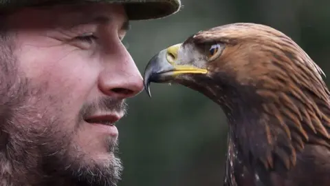 David Trenier, with a beard and flat cap, nose to beak with Loki the golden eagle. 