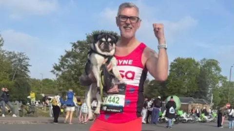 Wayne Goodhead Wayne has grey hair and black glasses and is wearing a pink vest with white writing that reads "Wayne, PDSA". His race bib is black with green numbers on it. He is holding a fist in the air and with the other hand is holding a small black and white pug