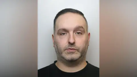 West Yorkshire Police A police mugshot of a man in his late thirties. He has short dark hair and stubble.