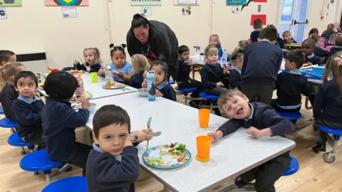 Young children are sat at tables eating lunch in a primary school canteen. Some are smiling at the camera. Kerrie, a parent who now works at the school, is bending down to talk to a young girl in the background. 