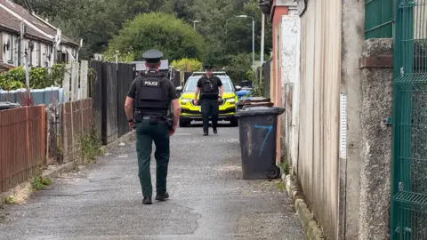 Pacemaker Two police officers standing in an narrow alleyway, which has a row of houses and fences to the left. There is a police car at the end of the alleyway. There are a few bins on the right up against a wall.