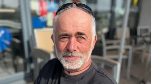 A man with short grey hair and sunglasses on his head and a grey chinstrap beard and moustache looking towards the camera. 