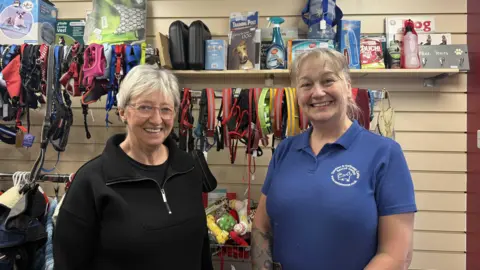 Two women, one with a black jumper, glasses and short hair, and another with a blue t-shirt saying Dumfries and Galloway Canine Rescue Centre, stand in front of a shop display of dog products.