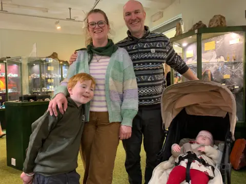 A young family at the Gem Rock museum, the youngest child is sound asleep in her buggy. A young boy with red hair is leaning into his mother who has glasses and her hair tied back. The father has a patterned jumper, a bald head and a beard.