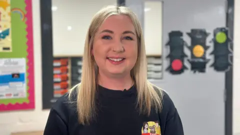 A woman with blonde hair stares into the camera. She is wearing a black top and has a nose ring. There are brightly coloured items on the wall behind her.