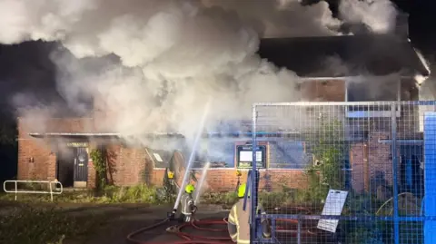 Thick plumes of smoke billowing from the a derelict building as firefighters aim water jets at the building. 