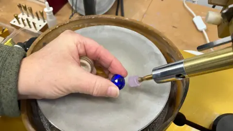 BBC Images A hand holding a deep-blue gem stone. There is a piece of equipment in vision which is being used to polish the stone. 