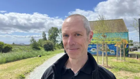 A man with very short grey hair smiling into the camera with black polo shirt an fleece on. Behind you can see a colourful mural depicting a river and sky and to the left is a footpath with a river and shrubbery to the left of it.