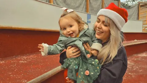 Katie Tradie Katie Tradie, holds her daughter in her arms, both of them smiling. Her baby daughter wears a green dress with gold stars and a blue raincoat. Katie wears a black hoodie, red santa hat and has long blonde hair. 