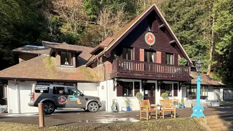 Isle of Man Farm Shop Company The modern version of the Swiss Chalet has a white painted ground floor with oak shutters on the windows. The first floor is pitched and is wooden panelled. There is a traditional balcony over the front of the building. Behind it is trees, and there is a 4x4 car parked outside to the left.