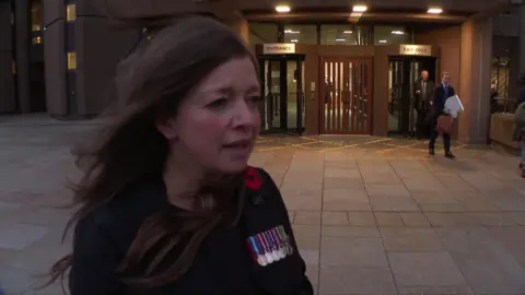 Elaine Overend, who has long brown hair and wears a blazer with a poppy badge and military medals pinned to it, talks to the camera outside Liverpool Crown Court, 