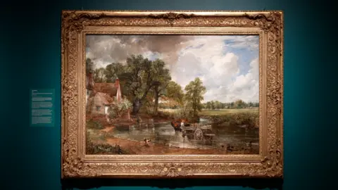 Qays Najm/BBC A large oil painting displayed on a wall as part of an art exhibition. The painting depicts a rural scene with a horse and cart moving through a river. There is a cottage on the bank of the river. 