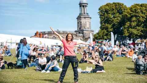 Beth Heath has shoulder-length brown hair and is wearing a pink t-shirt and black trousers. She has both arms out in the air. She is standing on green grass with dozens of people sitting and standing on the grass behind her. In the background is a church and white tents.