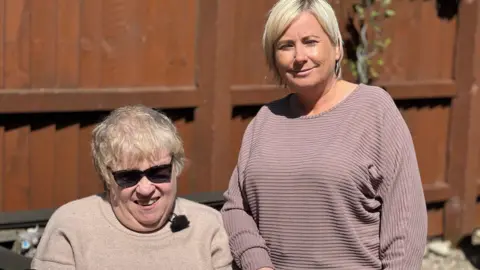 Resident Linda sitting down with cropped white/blonde hair and sunglasses while care support worker Donna stands next to her wearing a purple long-sleeved top and a blonde bob