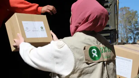 Ghiwa AbiHaidar/Oxfam A woman wearing a purple hijab and Oxfam top is handed a box from the back of a lorry.