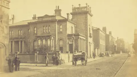Edwin Williams Sepia image of Winckley Square and Cross Street in Preston in 1862 showing an impressive large square two-storey building with a tower to the right, a low wall to the front and a cobbled street. There are groups of smartly-dressed men at the corners of the street and a horse and cart is going past to the right