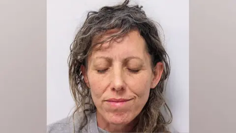 Police mug shot of Kara Alexander, a woman with brown and grey curly hair, with her eyes closed 