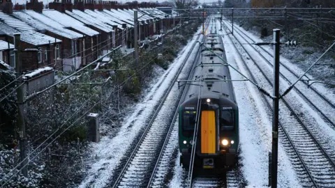 Library photograph of a black and yellow train as it travels on tracks covered in snow. Houses on the left of the tracks have snow on their roofs.