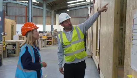 Sussex PCC A man in a grey jumper wearing yellow high-vis vest and white hard hard, while pointing to chipboard walls with his left hand. A woman wearing a dark blue top, light blue high-vis vest and orange hard hat is looking to where the man is pointing. They are stood in a large warehouse