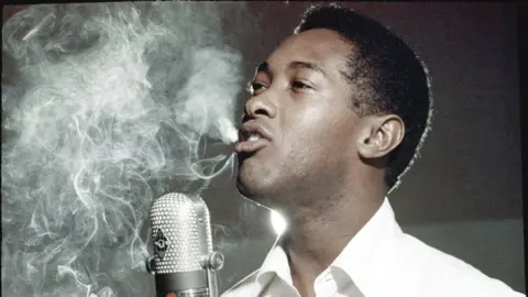 Getty Images Sam Cooke performs in a recording studio. A radio microphone is front of him, and smoke - probably from a cigarette - is visible in the air. He has dark hair and is wearing a white shirt, with the photo showing him between words in his song.