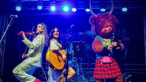 Tiree Music Festival Performers play violin and guitar under bright blue stage lights, while a large Highland cow mascot in a tartan kilt stands at the front of the stage holding a wind instrument.