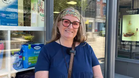 A person stands outside a pharmacy, wearing glasses and a patterned headband, with healthcare products displayed in the shop window behind them.