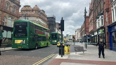 A street in Leeds with police tape across the road, two green double decker buses are parked up and there are several police cars in view. 