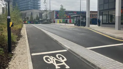 A clearly segregated cycle lane which is separated from the main road with a slim section of paving. It is a spacious scene with a bus stop on the side.