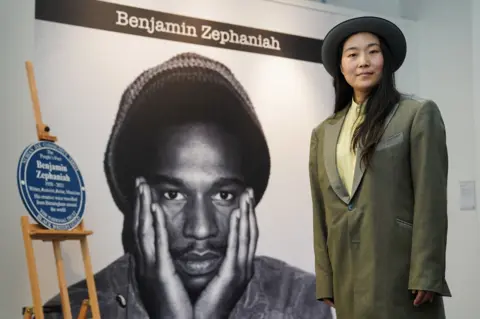 PA Media Qian Zephaniah wearing a brimmed hat and green and gray jacket in front of a portrait of Benjamin Zephaniah and a blue plaque at a recent event.