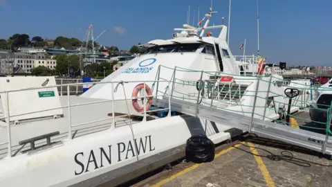 Islands Unlimited boat, San Pawl, a catamaran style vessel. There is a ramp leading on to the boat. In the background is housing.