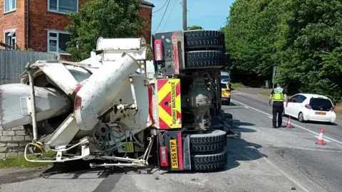 An overturned cement lorry has crashed into a brick wall and fence. Police officers are stopping traffic in the road next to it. The lorry is white with black tyres and red and yellow "highway maintenance" markings on the back. A house and trees can be seen to either side of the road.