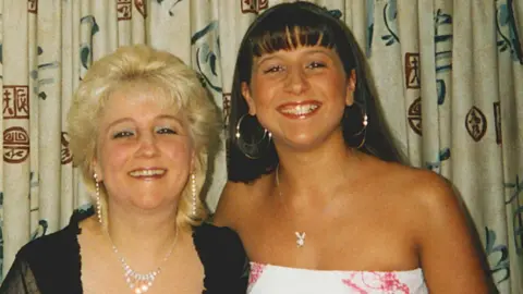 Family photo Pauline, in early middle age, and Jenna as a teenager. They are both dressed up. Pauline has layered, short blonde hair, is wearing makeup and diamante necklace and earrings. She has a black scoop neck top with ruffles at the edges and short, slightly see through lacy sleeves. Jenna has long dark hair and a fringe. She is tanned and wears a strapless white dress with red detailing either side. She has a number of very large hoop earrings and a silver chain with a pendant at the end. They are standing in front of curtains and both are smiling broadly.