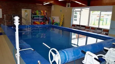 ST GEORGE'S HYDROTHERAPY POOL A small swimming pool filled with water. The pool has a blue cover, which is rolled back. There are chairs and shelves full of inflatables next to the pool.