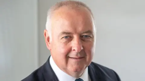 Staffordshire PFCC A close-up of a man's face, smiling at the camera. He has short grey hair and is wearing a white shirt under a dark suit jacket