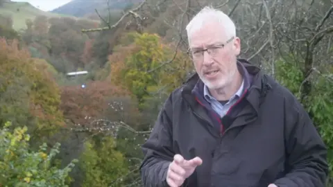 BBC Reporter Craig Duggan stands in a valley of autumn trees, the train carriage of one of the crashed trains is visible in the background. Craig Duggan is wearing a black raincoat and has grey hair and is wearing glasses. 