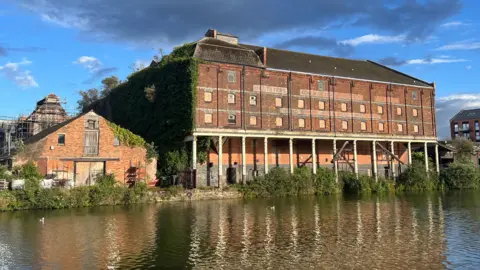 Downing's Malthouse at Baker's Quay in Gloucester. It is a large red brick building right on the waterfront, with pillars supporting the upper levels. It is very overgrown, with boarded up windows and fading signage.