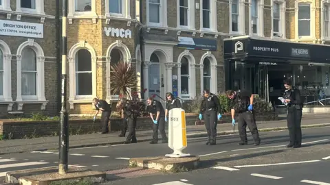 Matthew Ebers A line of police officers stood in the road. They are crouched over while searching the pavement and road surface.