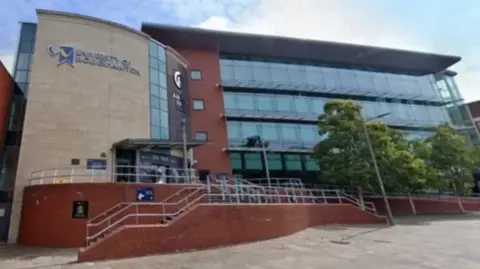The orange brick and glass building has stairs leading to it on the left and right, in front of it. There are some tall tree on the right shielding some of the glass near to some lamp posts.