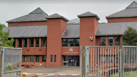 A red brick building with a black roof, there is the courthouse symbol above two blue doors. The image is taken behind the silver metal gates, a red and white barrier is in-view.