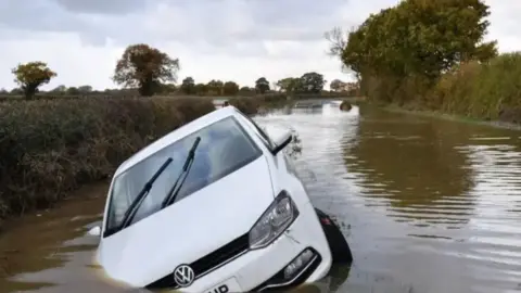 A white VW is partially submerged in floodwater - the water on the road resembles a river.